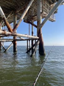 View of a fishing platform with rusted supports, showcasing a fishing rod in the foreground and a boat in the distance.