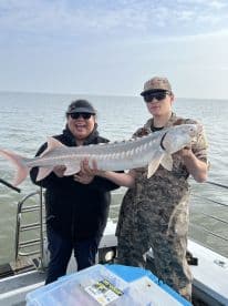 Two anglers proudly displaying a large Sturgeon on a fishing charter, with the open water in the background.