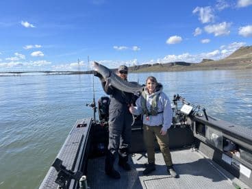 Two anglers proudly displaying a large Sturgeon while fishing on a scenic river, showcasing the beautiful surroundings and clear blue skies.