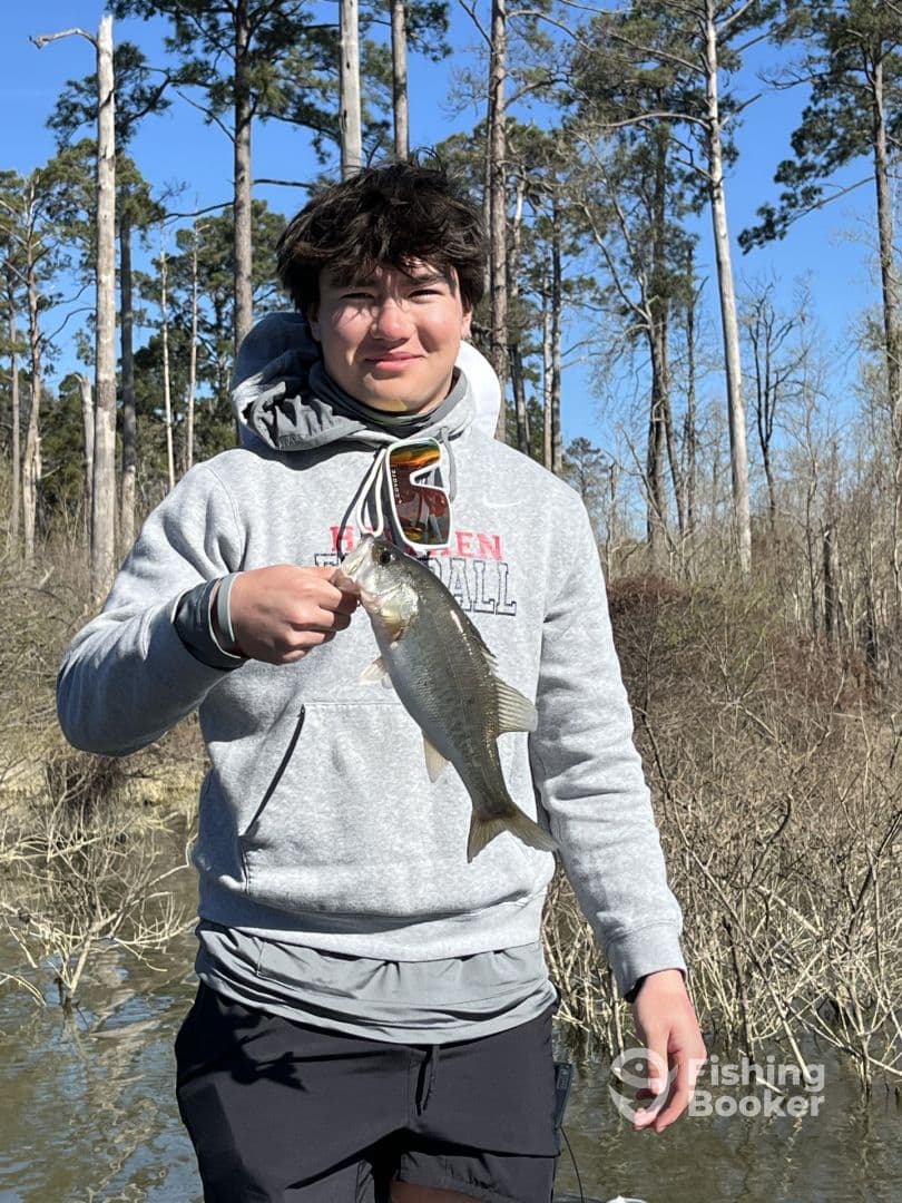 A young angler proudly displaying a Largemouth Bass while fishing in a scenic freshwater environment surrounded by trees.