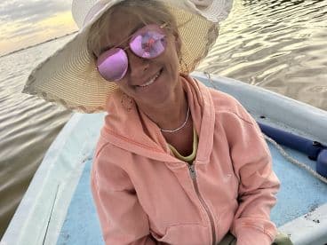 A smiling woman wearing a large sun hat and sunglasses sits comfortably in a small boat, enjoying a peaceful moment on the water.