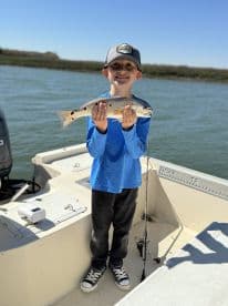 A young angler proudly displaying a Speckled Trout while fishing on a boat, enjoying a sunny day on the water.