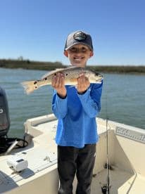 A young angler proudly displaying a Speckled Trout while fishing on a boat, showcasing a sunny day on the water.