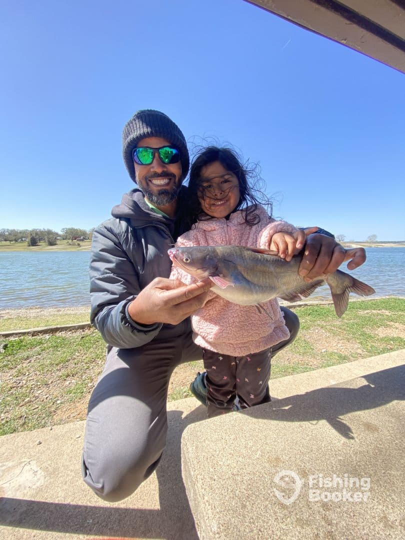 A proud moment as a young girl holds a sizable catfish alongside an adult at a scenic fishing spot, showcasing a successful catch.