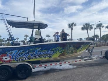 A young angler and an adult proudly display their catch on the 'Holy Smokes' charter boat, set against a backdrop of palm trees and a sunny sky.