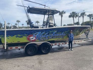 A young angler poses beside the 'Holy Smokes Fishing Charters' boat, showcasing its vibrant design in a sunny marina setting.