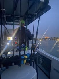 A view from the helm of a fishing boat at dusk, showcasing fishing rods and electronic equipment, with a city skyline illuminated in the background.
