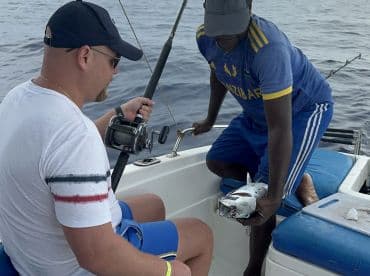 A fisherman actively reeling in a fish while another angler assists on a boat in open waters.