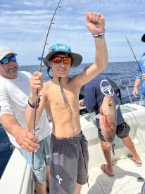 A young angler proudly displaying a Snapper while fishing on a boat, surrounded by fellow anglers in a sunny offshore setting.
