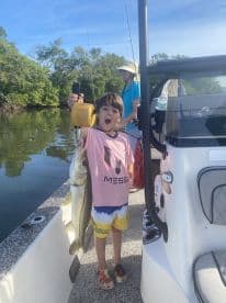A young angler excitedly holds up a large Snook while fishing on a boat in a lush mangrove setting.