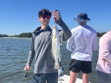 A young angler proudly displaying a Speckled Trout while fishing on a sunny day, with fellow anglers in the background.