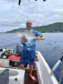 An angler proudly displaying a large Trevally while fishing from a boat in a tropical setting.