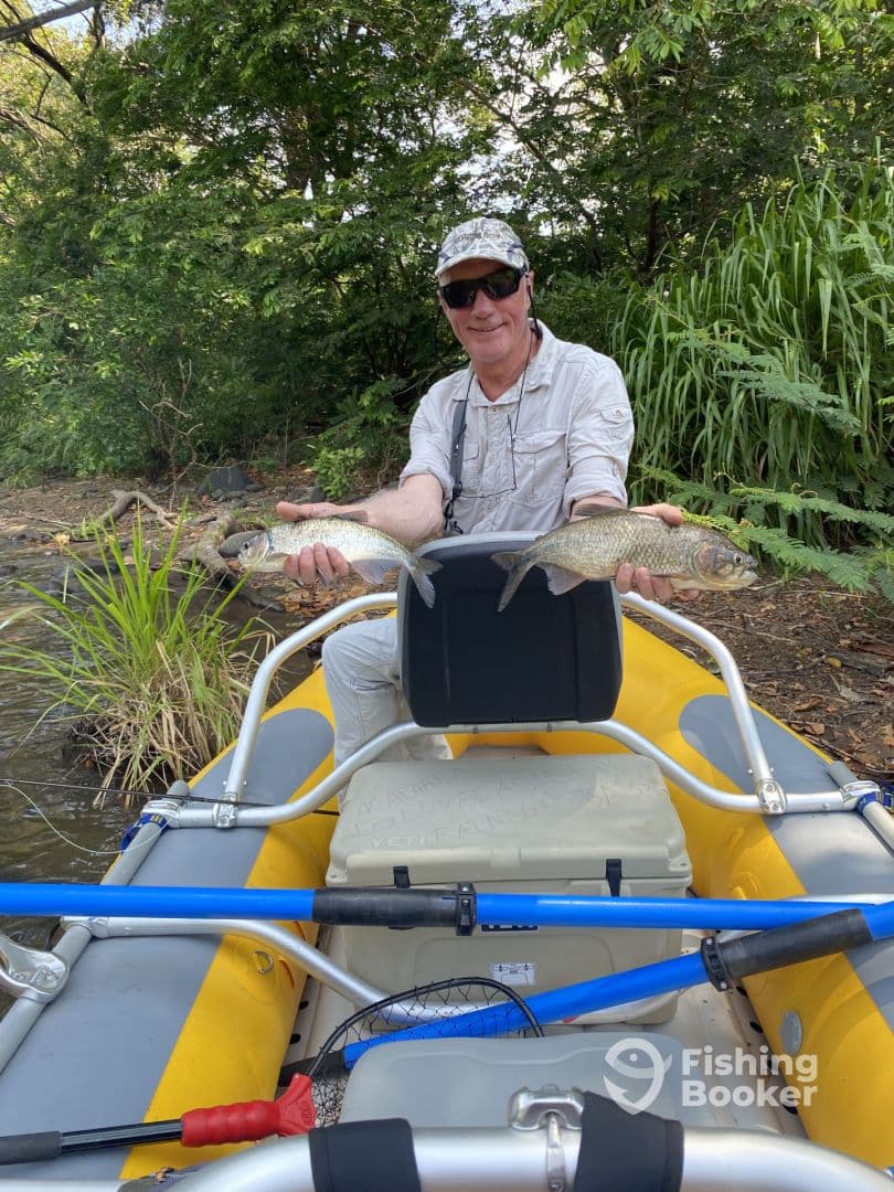 An angler proudly displaying two freshly caught fish while seated in an inflatable kayak amidst a lush, green environment.