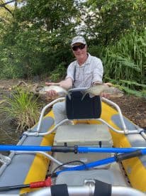 An angler proudly displaying two freshly caught fish while seated in an inflatable kayak amidst a lush, green environment.