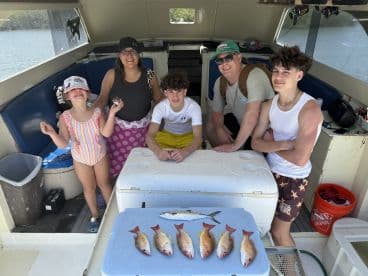 A family of four proudly displaying their catch of Snapper and a Mackerel on a fishing charter, enjoying a sunny day on the water.