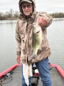 An angler proudly displaying a large Largemouth Bass while fishing from a boat on a calm lake.
