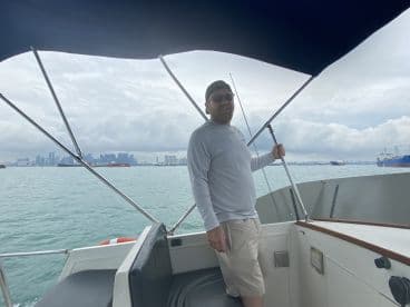 A smiling angler at the helm of a boat, navigating through a scenic waterway with a city skyline in the background.