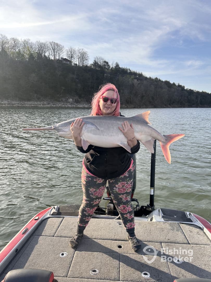 An angler proudly displaying a large Sturgeon while standing on the deck of a fishing boat, surrounded by a scenic lake backdrop.