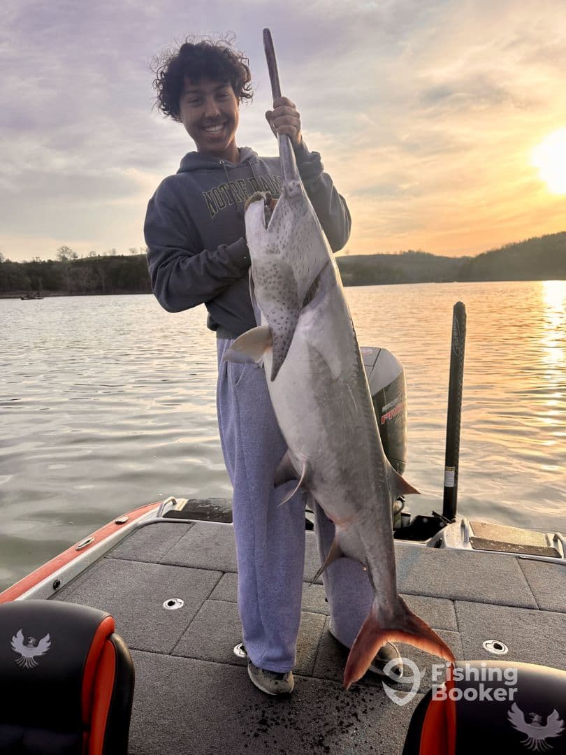 A young angler proudly displaying a large catfish while standing on a fishing boat at sunset, showcasing a successful day on the water.