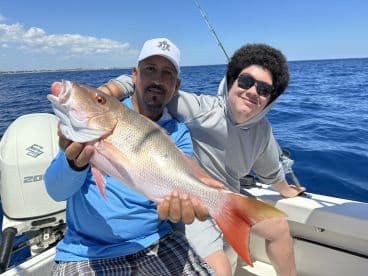A proud angler showcasing a vibrant Snapper while fishing on a sunny day in open waters, accompanied by a young enthusiast.