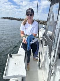 An angler proudly displaying a freshly caught fish while aboard a fishing boat, with a scenic backdrop of the water and rocky jetty.
