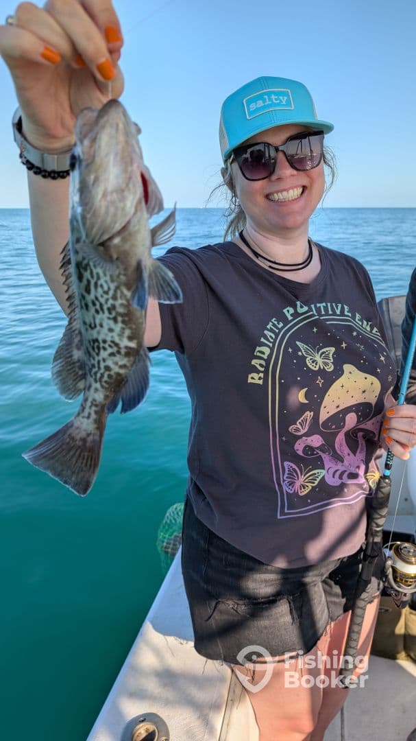 A smiling angler proudly displaying a Spotted Grouper while fishing on a sunny day in clear waters.