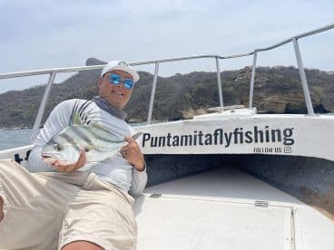 Angler displaying a vibrant Roosterfish while seated on a boat, showcasing a scenic coastal backdrop.