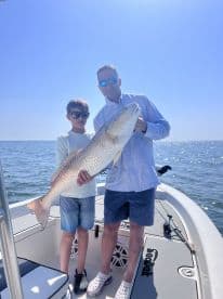 A proud father and son duo holding a large Redfish while fishing on a sunny day, showcasing their catch on a boat.