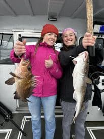 Two anglers proudly displaying their catches, a large Rockfish and a Cod, aboard a fishing charter in a coastal setting.