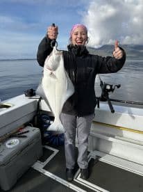 A young woman proudly displaying a large Halibut while fishing on a boat in a scenic coastal environment.