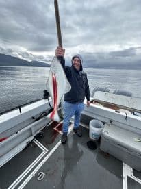 Angler proudly displaying a large Halibut on a fishing boat, showcasing a successful catch in a scenic marine environment.