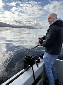 An angler reeling in a catch while fishing on a calm day, surrounded by scenic mountains and a clear sky.