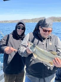 Two anglers celebrating a successful catch of a Lake Trout while fishing on a boat, showcasing the beautiful lake scenery.