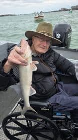 An elderly angler proudly displaying a freshly caught fish while seated in a wheelchair on a fishing boat.