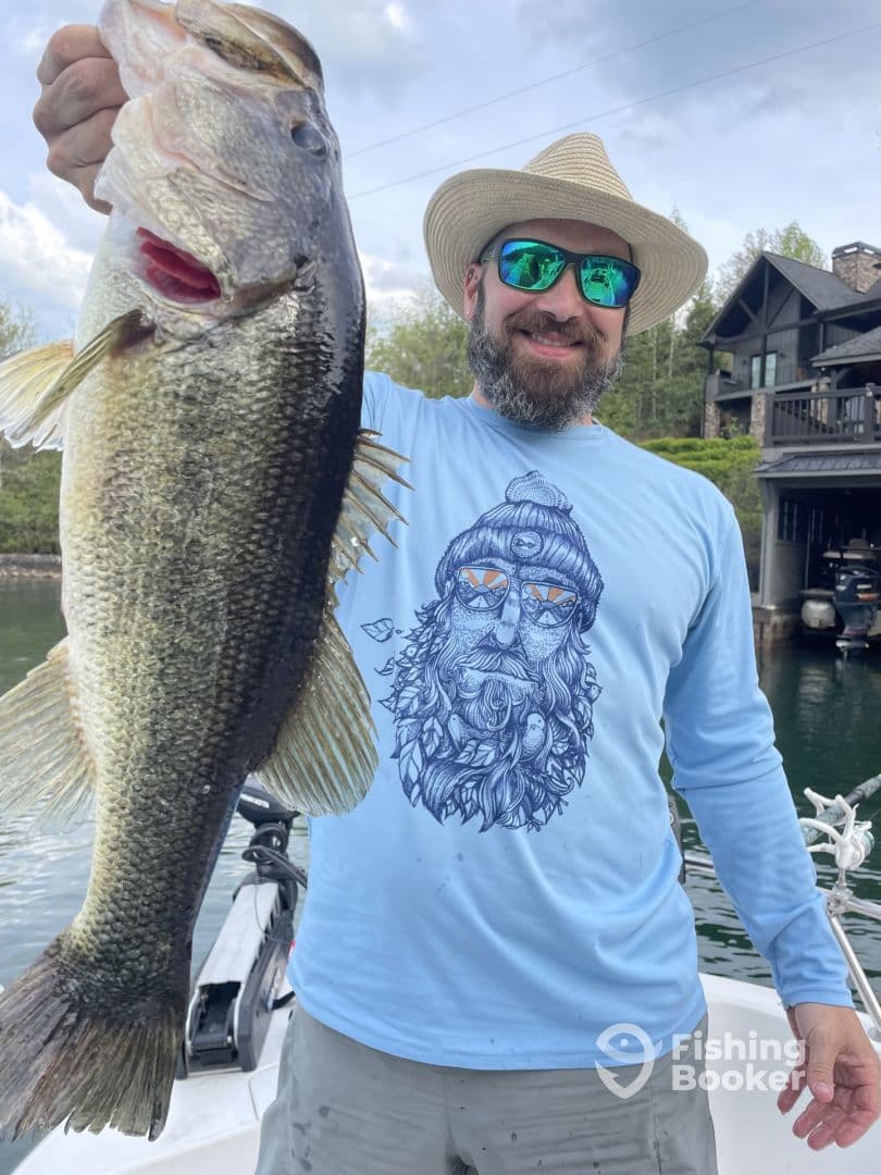 An angler proudly displaying a large Largemouth Bass while fishing from a boat, with a scenic lakeside home in the background.