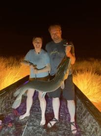 Two anglers proudly displaying a large Northern Pike on a boat during a night fishing trip.