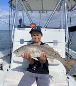 A young angler proudly displaying a large Redfish while seated on a boat, showcasing a successful fishing trip.