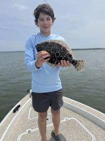 A young angler proudly displaying a Flounder while standing on the bow of a boat during a fishing trip.