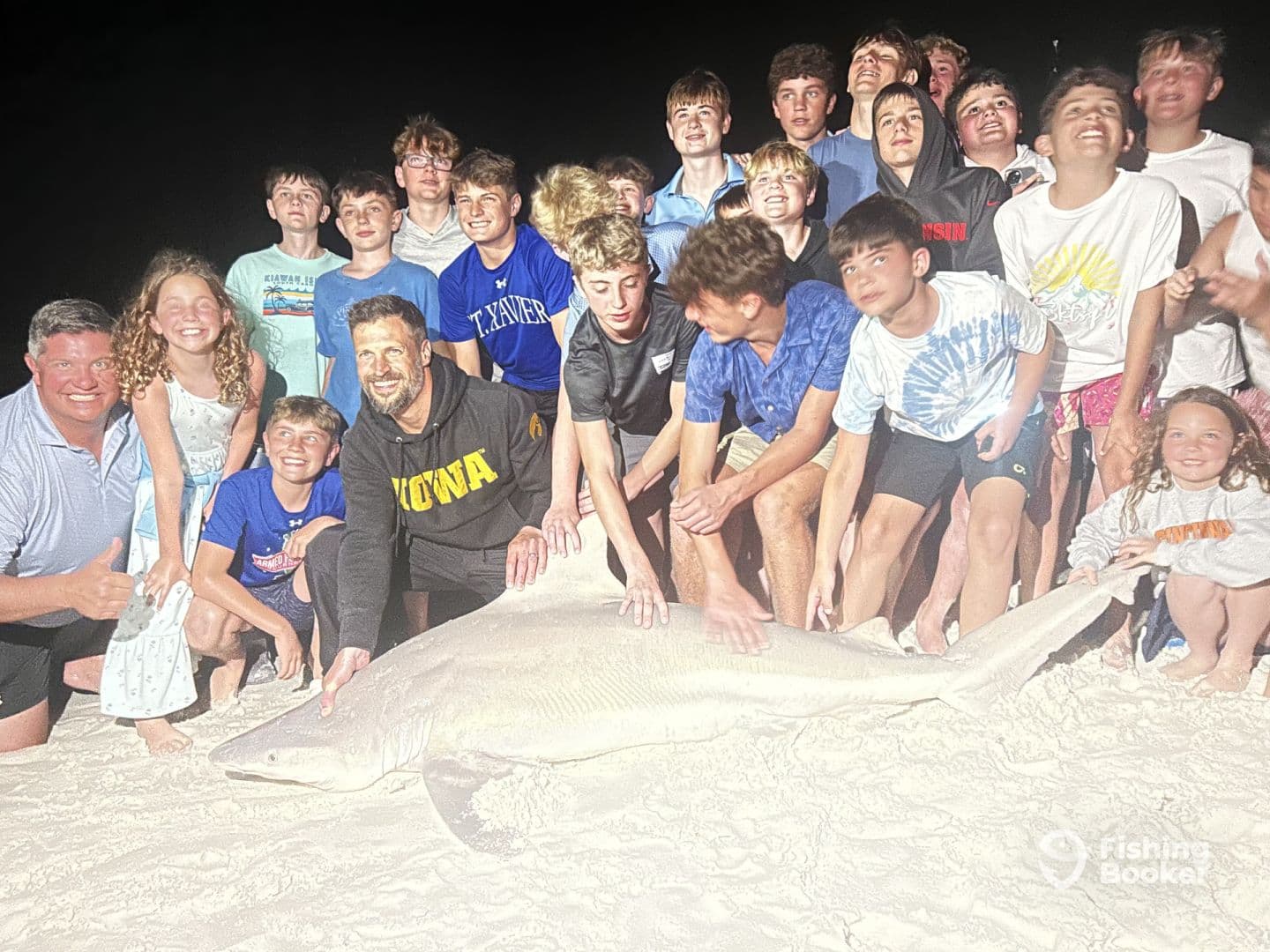A large shark is displayed on the beach with a group of excited young anglers and adults celebrating their catch during a night fishing trip.