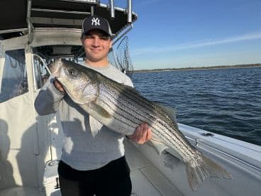 A young angler proudly displaying a large Striped Bass aboard a fishing boat, showcasing a successful day on the water.