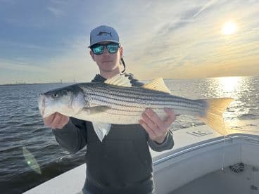A young angler proudly displaying a large Striped Bass while fishing on a scenic waterway at sunset.
