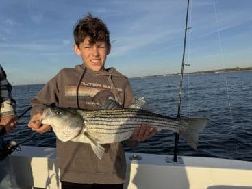 A young angler proudly displaying a large Striped Bass while fishing on a boat, showcasing a successful day on the water.