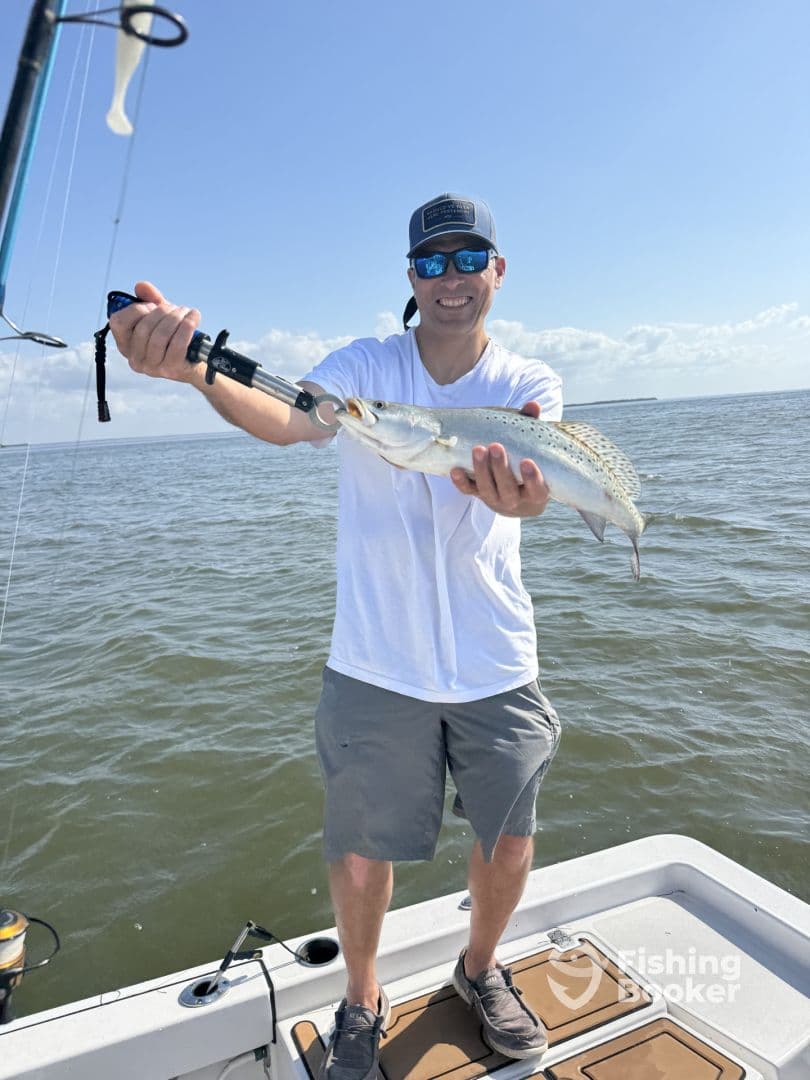 Angler proudly displaying a Speckled Trout while fishing on a boat in a sunny coastal environment.