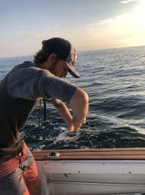 A young angler is actively reeling in a fish while fishing on a boat during a sunset, showcasing the excitement of the catch.