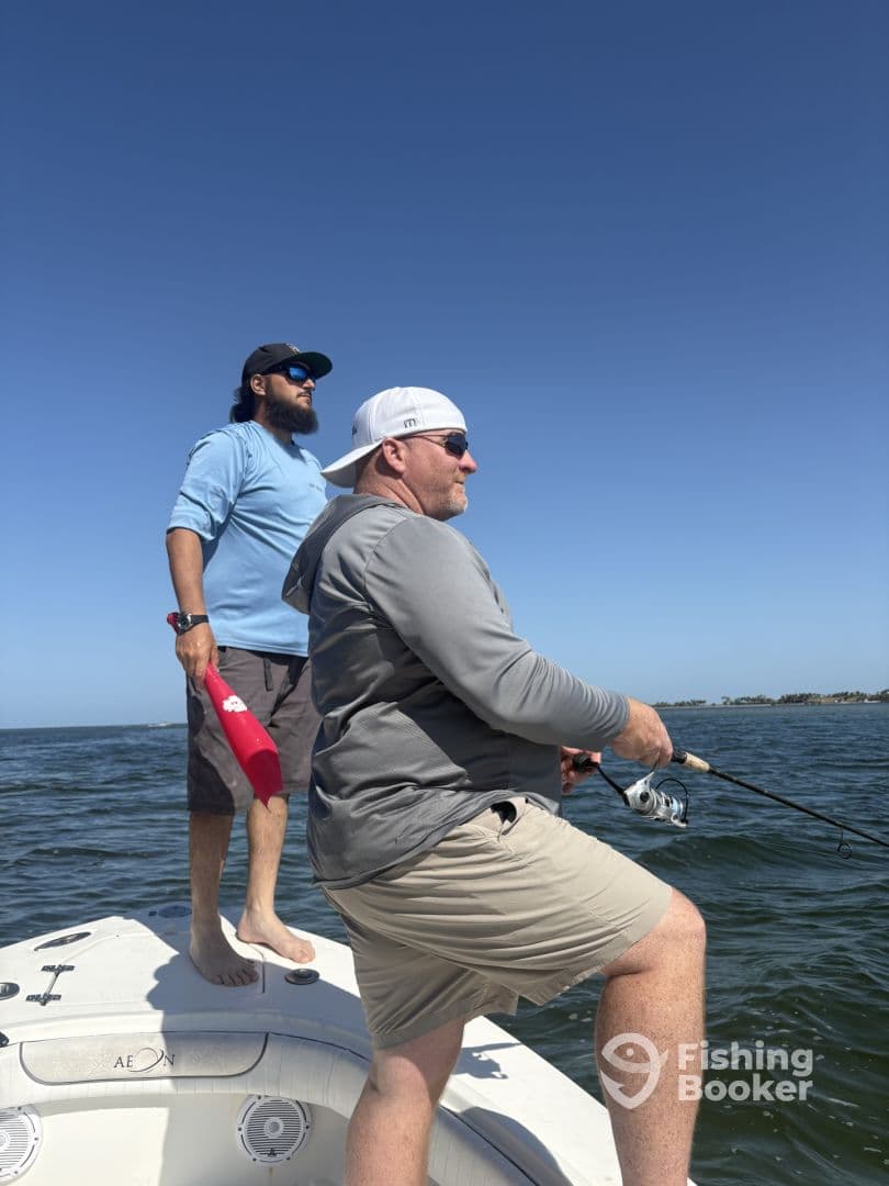 Two anglers actively fishing from a boat, with one reeling in and the other observing, set against a clear blue sky.