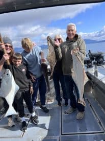 A family proudly displaying their catch of halibut and other fish aboard a fishing boat, enjoying a day on the water.