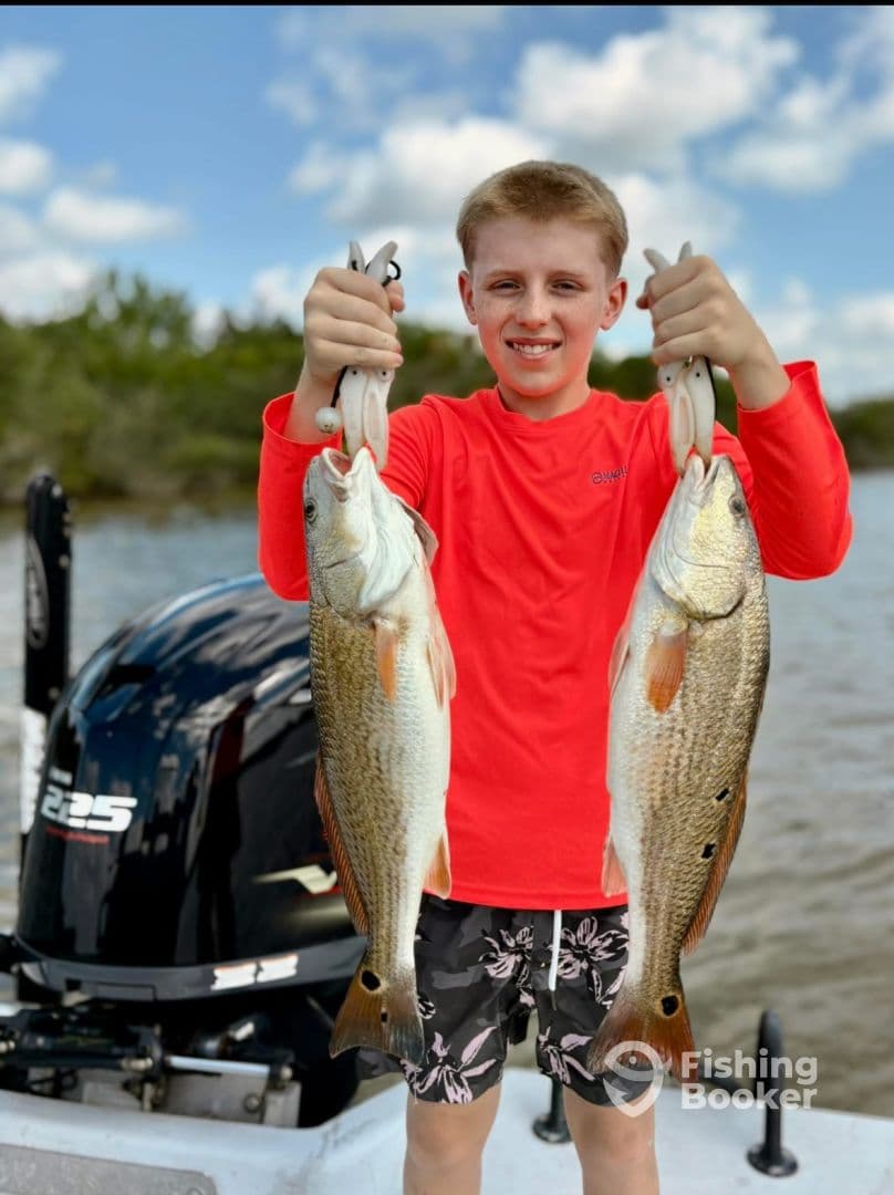 Young angler proudly holding two large Redfish while on a fishing trip, showcasing a successful catch against a scenic backdrop.