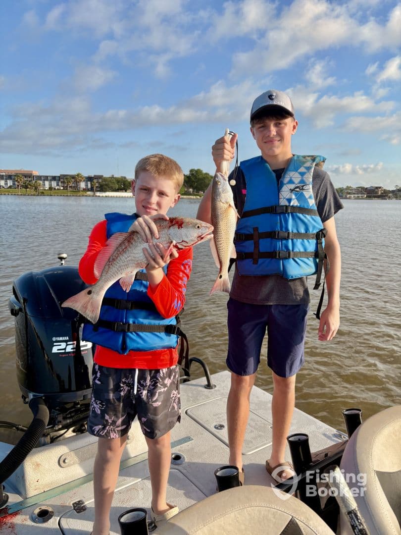 Two young anglers proudly displaying their catch of Redfish while on a boat, enjoying a day of fishing in calm waters.