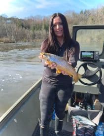 An angler proudly displaying a beautiful Brown Trout while fishing from a boat in a scenic river setting.