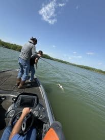 Two anglers actively reeling in a fish from a bass boat on a sunny day, showcasing the excitement of fishing in a freshwater lake.